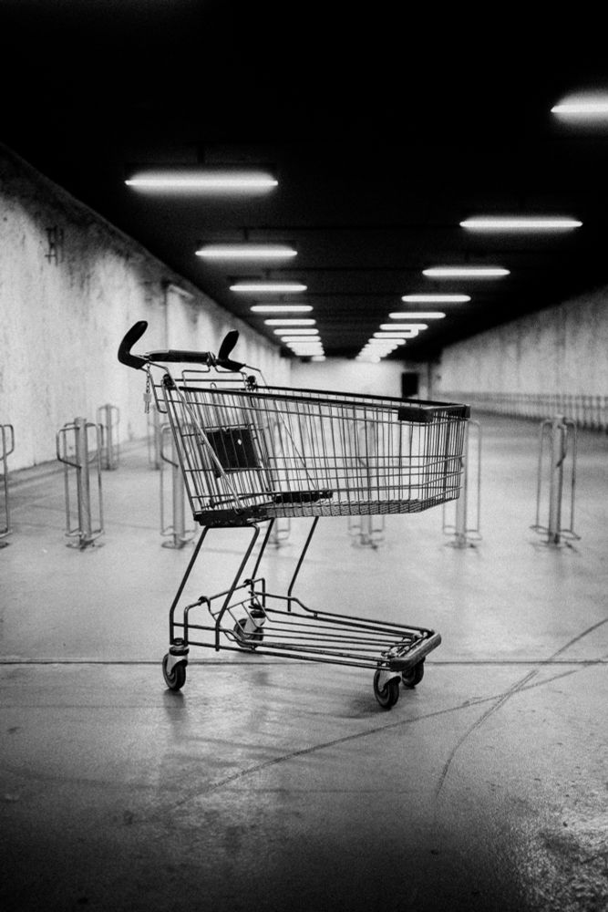 A black and white photo of an empty shopping cart standing alone in an underground or indoor parking area. The setting is stark and industrial, with rows of ceiling lights creating a vanishing point effect. The mood is quiet and slightly surreal.