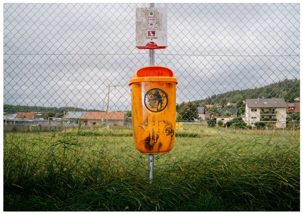 An orange public dog waste bin mounted on a pole in front of a chain-link fence, with grassy fields, rooftops, and forested hills under a cloudy sky in the background.