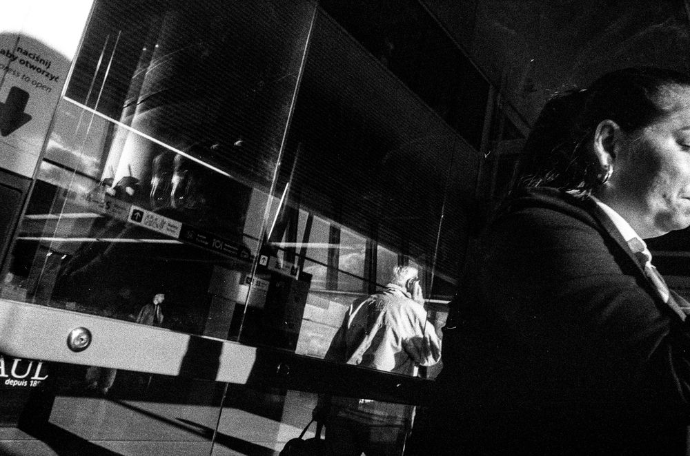 Black and white photo showing a busy urban scene with reflections on glass, a man walking in the background, and a woman in the foreground, creating layers of movement and depth.