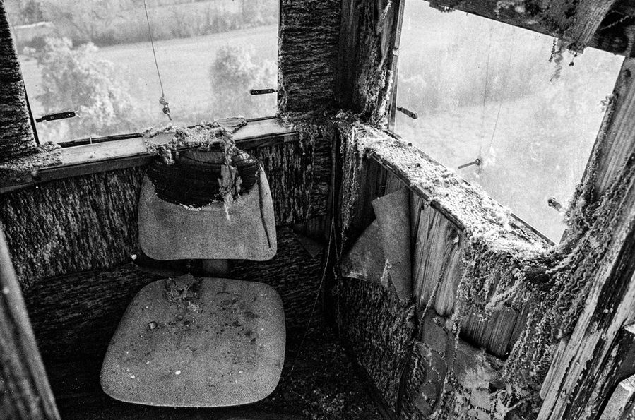 Black and white photograph of the interior of an old, decaying watchtower with a tattered chair and weathered wooden walls, overlooking a blurred landscape through broken windows.