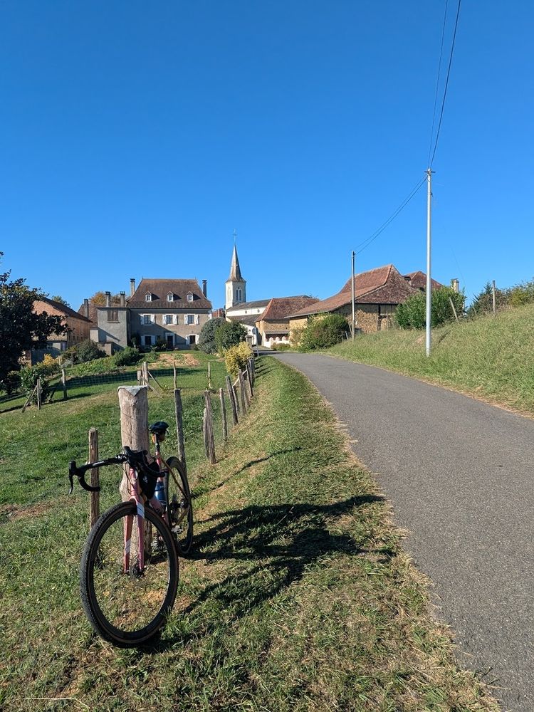 Même vélo, mais angle de vue différent, on regarde désormais derrière. Un petit bourg domine la route, on aperçoit un clocher et quelques fermes autour.