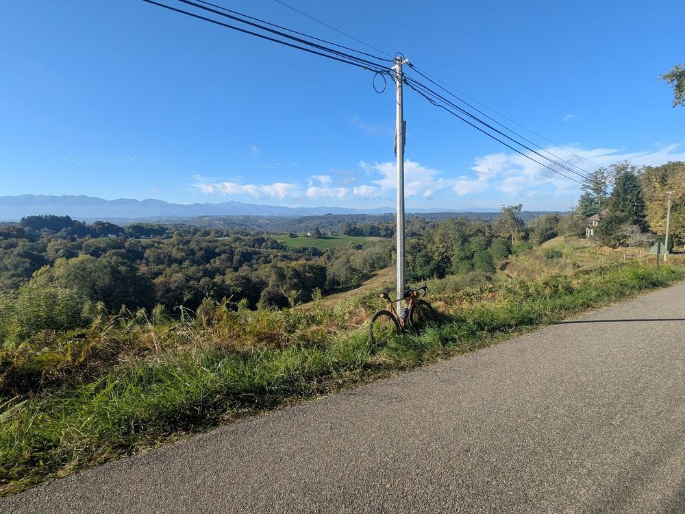 route surplombant une vallée, des collines boisées se trouvent derrière, et au fond les Pyrénées se détachent sur le ciel bleu.

Le vélo rose est appuyé contre un poteau.