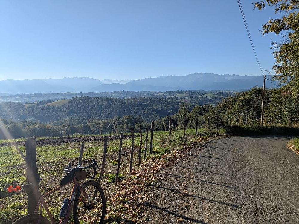 Sur la droite de la photo, une petite route communale qui descend. A gauche mon vélo posé sur un piquet de cloture délimitant un champs, des rayons de soleils illuminent le vélo, et font ressortir ses reflets dorés. Au fond on voit des coteaux couvert de forêts, et encore plus loin les Pyrénées. Ciel bleu dégagé.