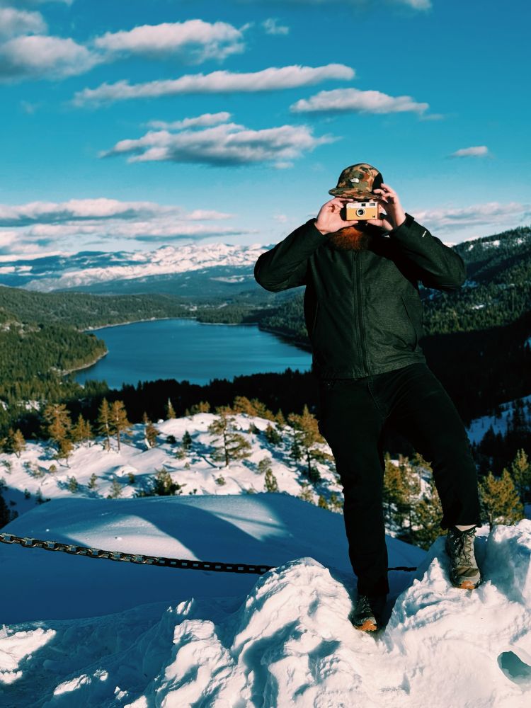 man standing in snow with hiking boots a coat and a hat holding a small film camera in front of his face. donner lake is in the background as well as the snowy sierras 