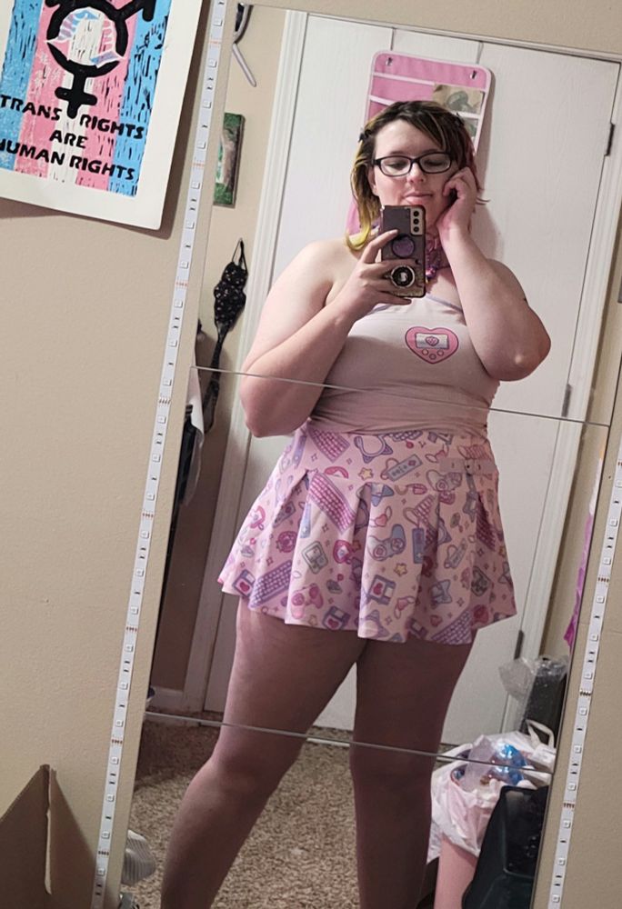 Echo taking a full body mirror selfy, she is wearing a light pink halter top and a short pink pleated skirt with a kawaii gaming print on it.