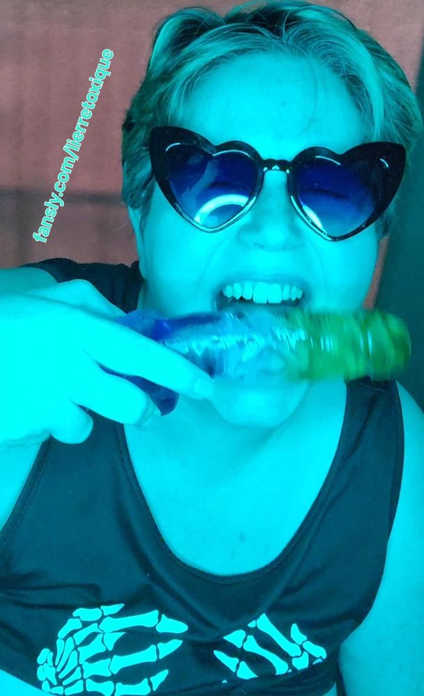 Lierre is a pale-skinned woman with short blonde hair. she is wearing a black tank with skeleton hands on the chest & black heart-shaped sunglasses. she is holding a blue to green ombre silicone dildo shaped like a human penis next to her face, and she is giving a toothy grin as if she's about to bite it