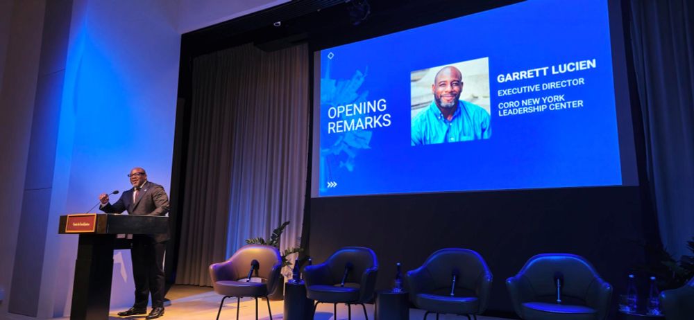 Garrett Lucien, Executive Director of Coro New York Leadership Center, stands at a podium delivering opening remarks at a leadership event. A large screen beside him displays his photo, name, title, and organization against a blue background. Three chairs and microphones are arranged onstage for a panel discussion.