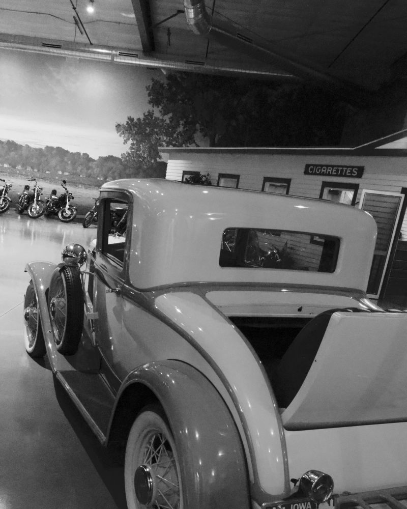 A timeless coupe from the early days of American motoring sits in quiet reverence under museum lights. Its polished curves and rumble seat recall an era when travel was an adventure, and every mile carried the promise of discovery. Behind it, a row of classic motorcycles and an old roadside shop sign complete the nostalgic scene — a tribute to the enduring spirit of the open road.
