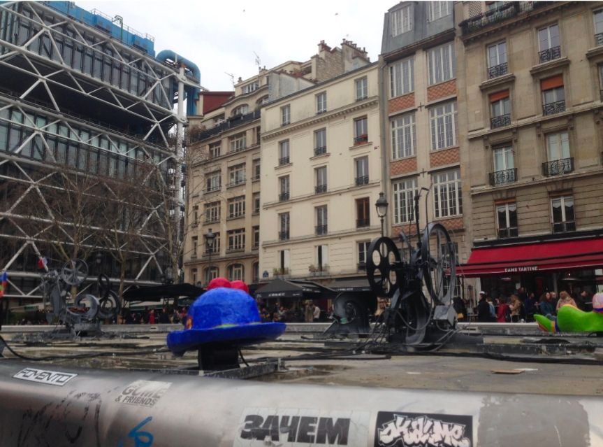 Brunnen mit bunten Figuren, wobei in dem Brunnen kein Wasser vorhanden ist.
Im Hintergrund eine Häuserzeile und links das futuristische Gebäude des Centre Pompidou in Paris.