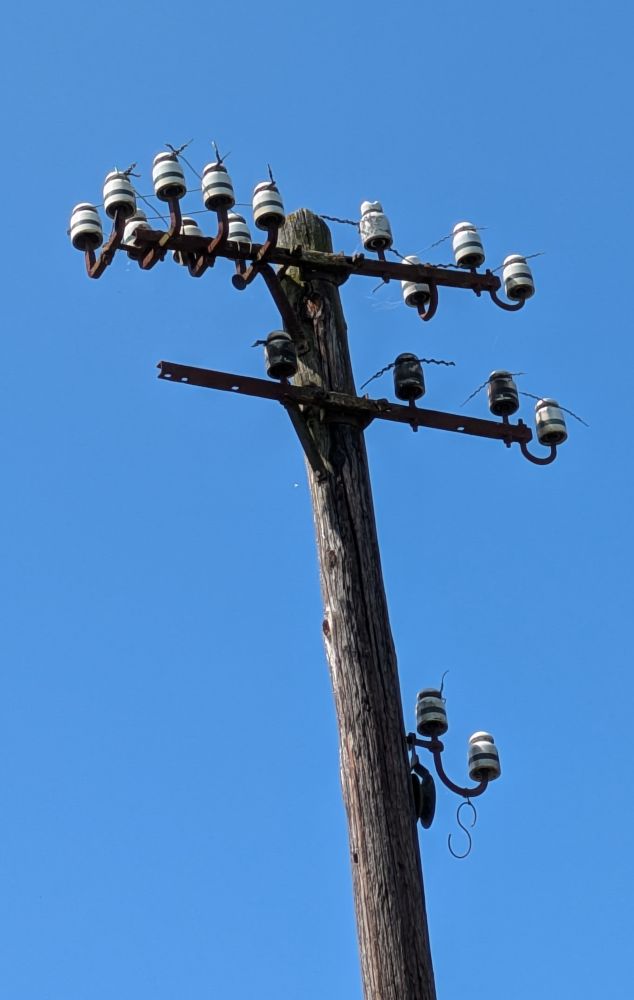 Alter Telegraphen Mast aus Holz. Oben an dem Mast sind zwei Querträger an denen mehrere Porzellan Isolatoren befestigt sind. Die Telegraphendrähte sind alle gekappt.