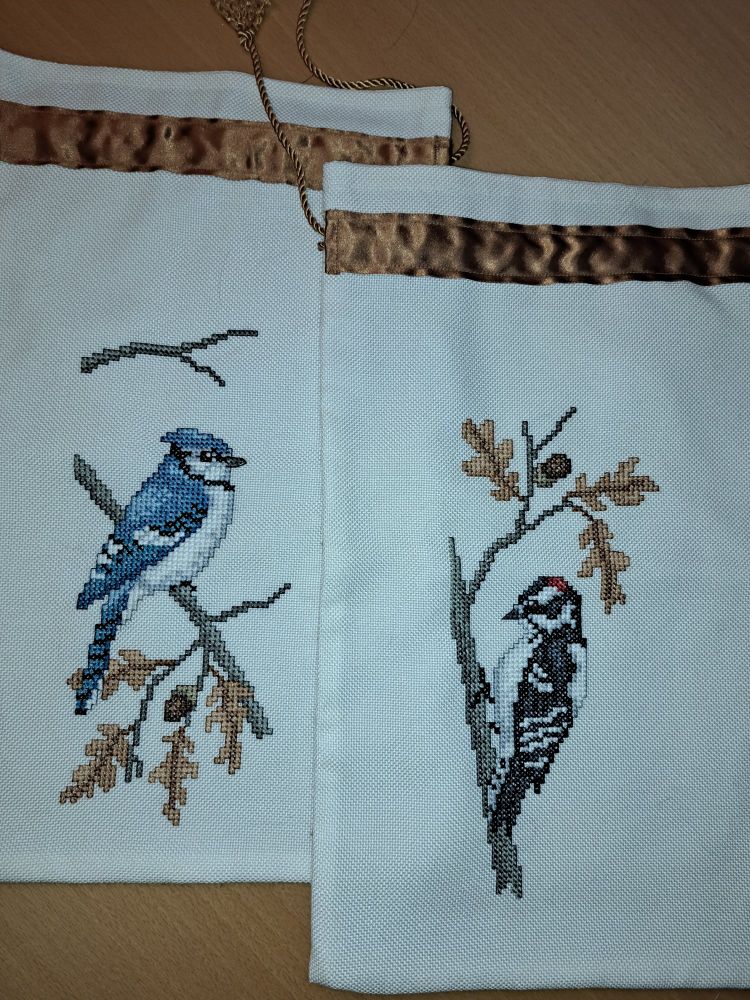 2 Fabric bags with cross-stitched birds. One is a blue Jay, the other is a downy woodpecker. Patterns by Paula Minkebige.