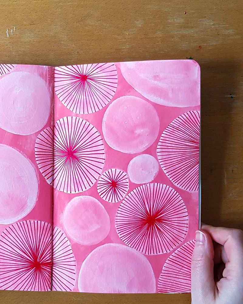 A hand holding the right hand side of an open sketchbook. The sketchbook spread is painted in bright pink with white translucent circles of varying sizes on top. Some of the circles have dark pink lines radiating out from the centre, similar to the spokes on a bicycle wheel.