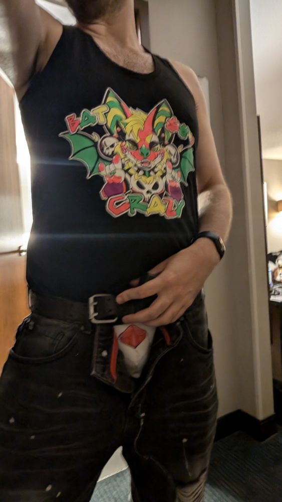 A person in a tank top with their pants unzipped to reveal a jock with a sheathed canine penis on it with the tip of the lipstick poking out just begging to be applied