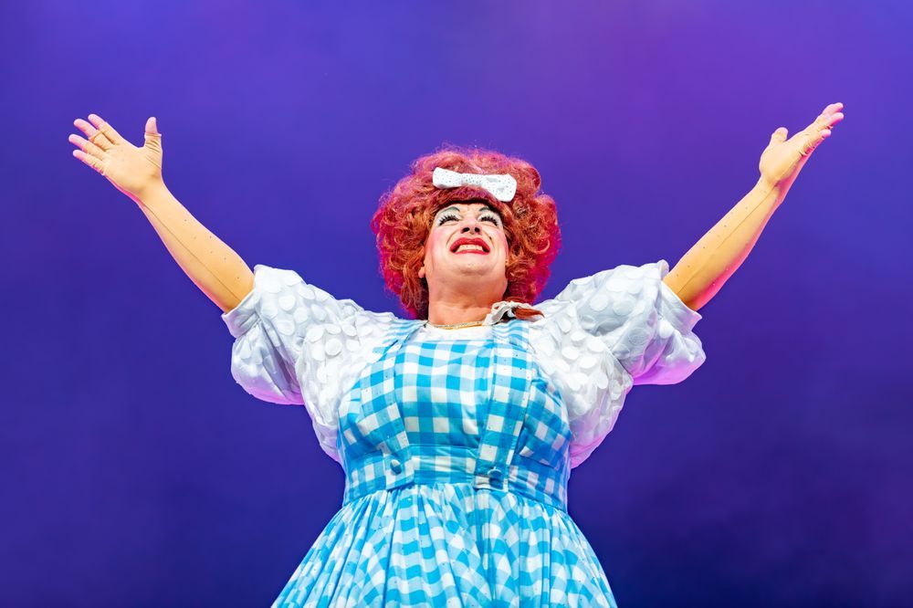 A panto dame with bright orange hair and a gingham dress is holding her hands up high and smiling.