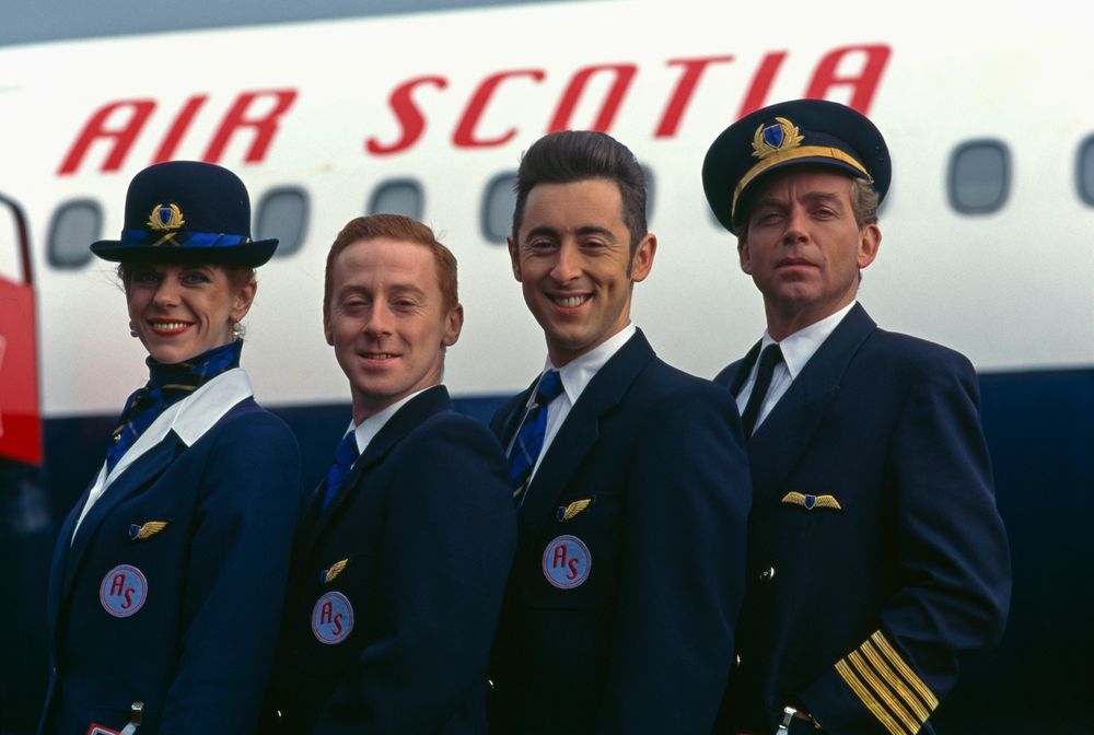 Four people in airline uniforms stand in front of a plane that reads AIR SCOTIA, smiling at the camera. 