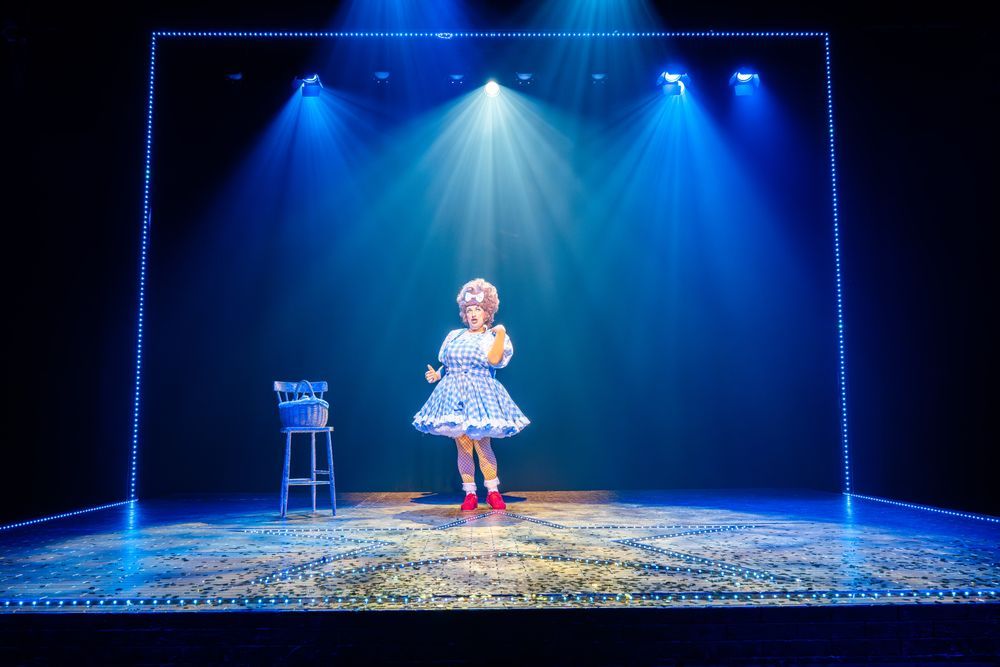 A panto dame stands towards the back of a stage where she is posing with her thumb up.