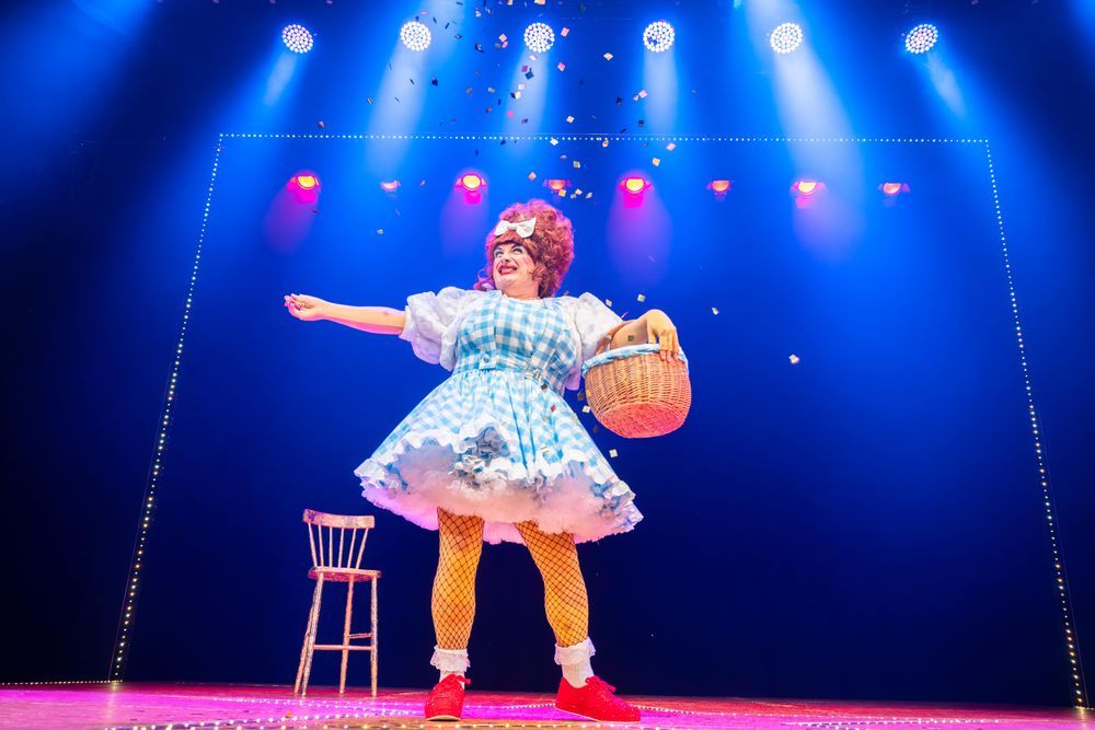A panto dame wearing a gingham dress and red trainers is throwing confetti out of a basket she has under her arm.