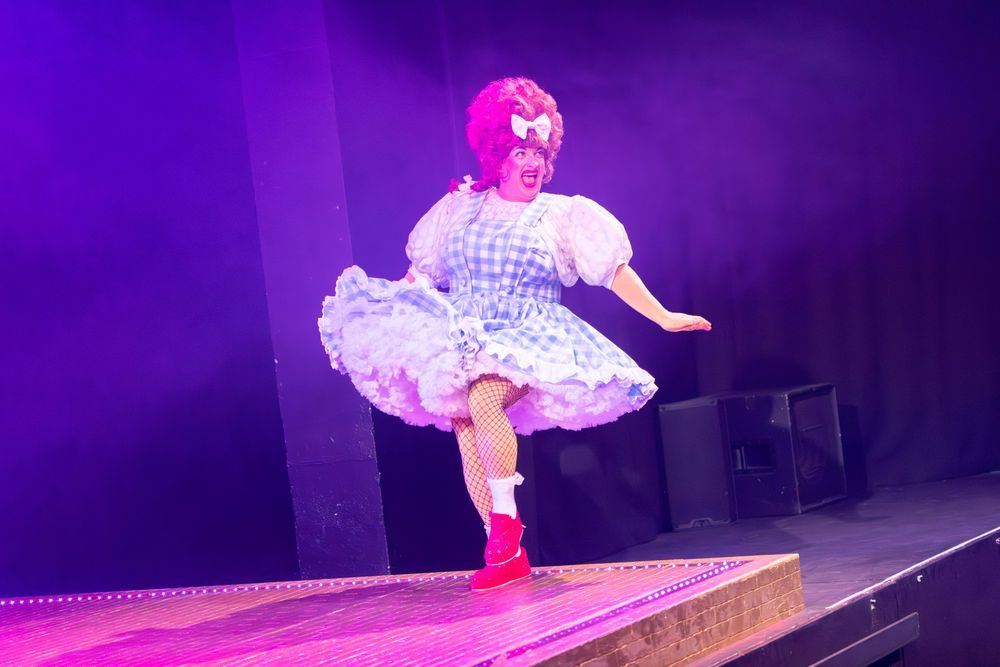 A panto dame in a gingham dress is skipping across the edge of a stage.