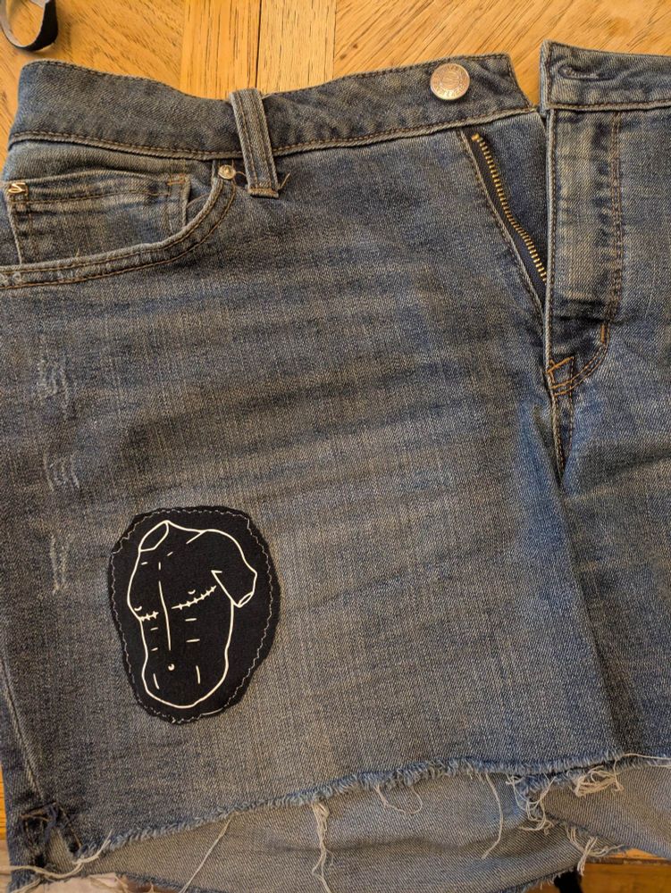 A medium blue pair of jean shorts with a rough hem patched with a dark blue denim patch. The patch has a white drawing of a trans guys chest with scars under the nipples