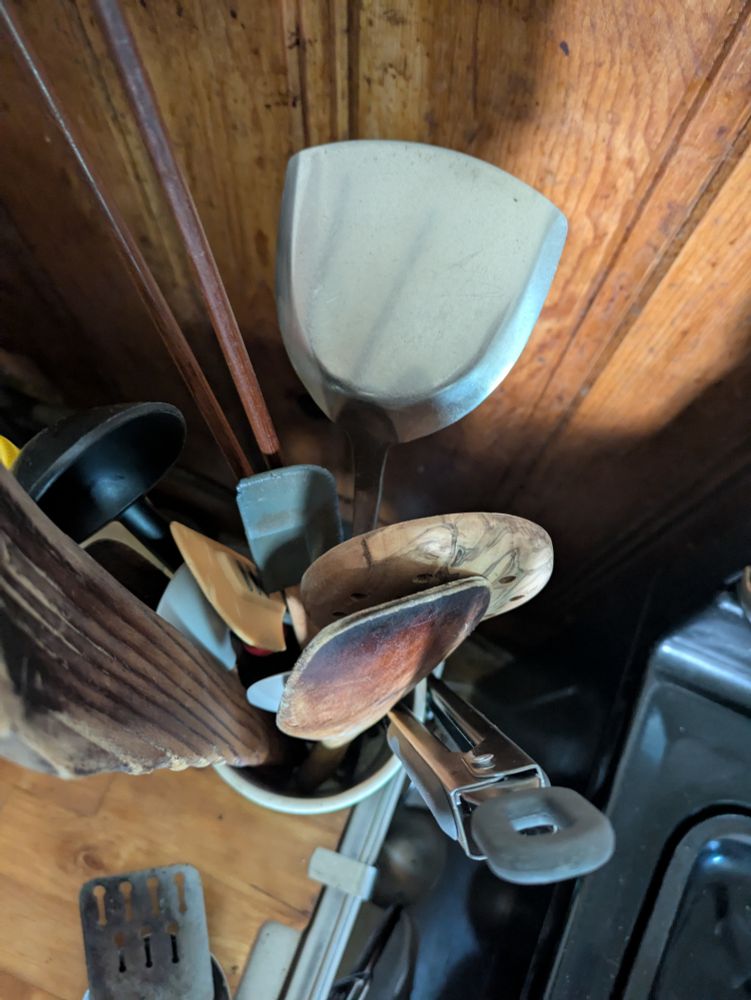 A top down view of a spatula jar. A mix of handmade wooden ones with metal, silicone and even one plastic spoon. Plus two cooking chopsticks