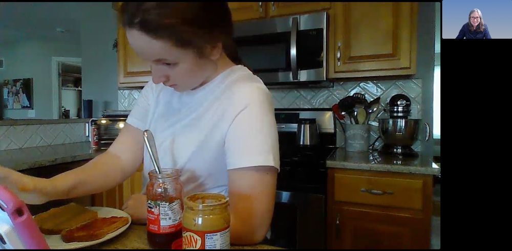 Tele-AAC session still shot with child touching her speech generating device.  Open jars of peanut butter and jelly are in front of her and there are two pieces of bread on a plate.
