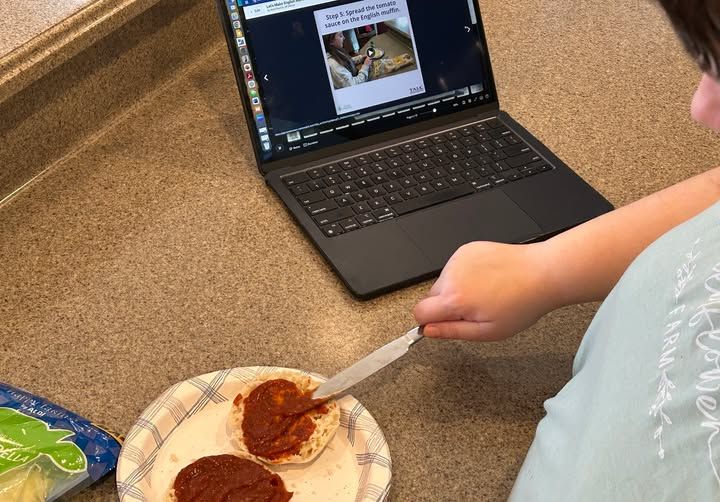 Person holding a knife and spreading tomato sauce on an English muffin. A laptop with a pizza recipe is open on the counter. 