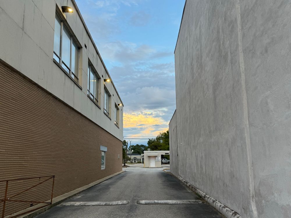 View down an alley with brick on one side, stucco on the other. It opens into a parki lot with a small kiosk building in the middle. 