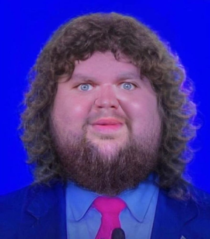 A digitally altered image of US VP JD “Just Dance” Vance wherein his face has expanded and he has shoulder-length curly hair. He is in front of a blue background in a blue suit and staring manically into the camera with a soft smile. 