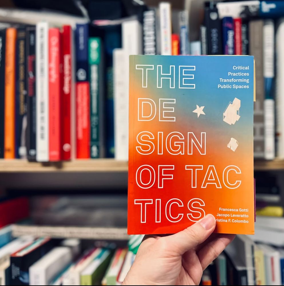 Photo of the book The Design of Tactics in front of a bookshelf. A hand is holding the book that has a colourful cover with gradients from blue (above) to red (below). The text on the cover is in big capital letters.