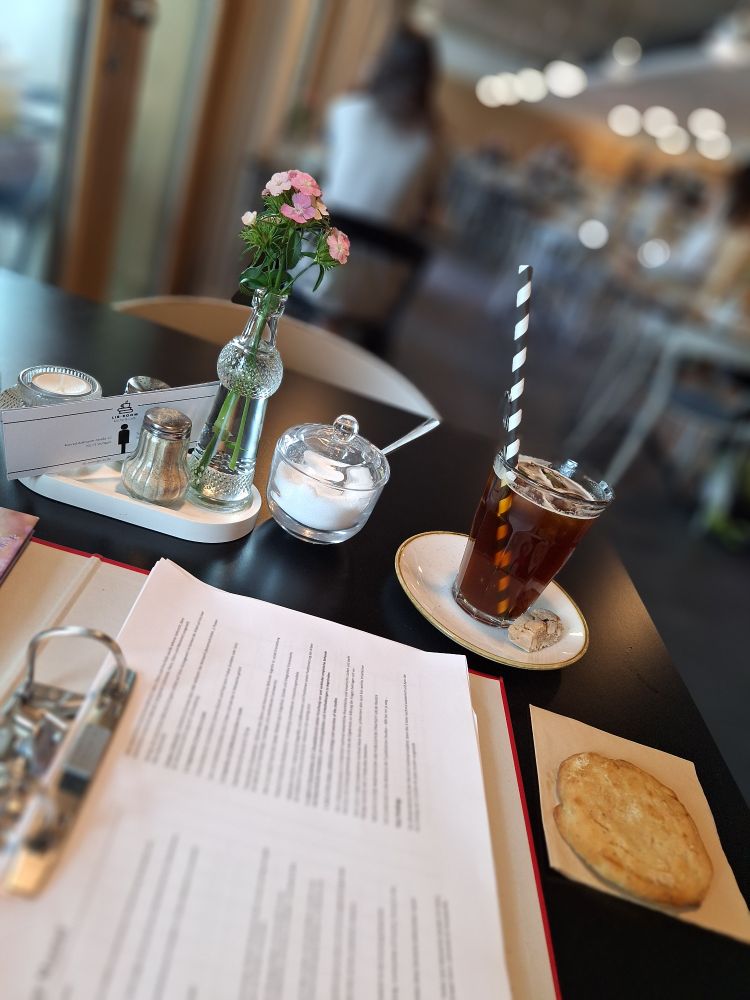 Ein aufgeschlagener Ordner mit einem Kaffee und Keks im Vordergrund vor einem unscharfen Café im Hintergrund 