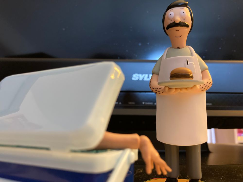 Bob of Bob's Burgers standing over and near a cooler. There's a human hand and forearm sticking out of it. Perfectly normal I mean who wouldn't want to try something new huh?