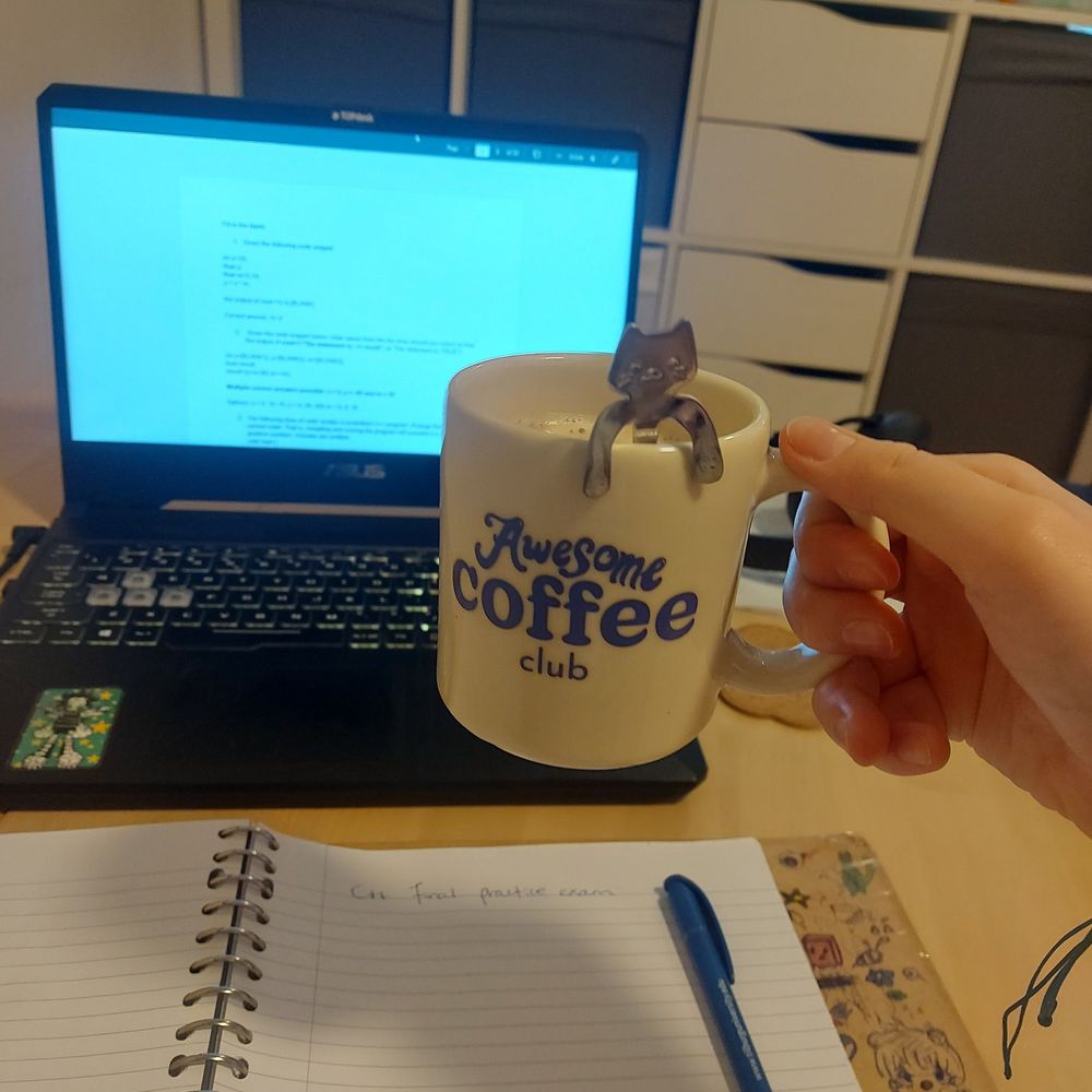 A picture of someone holding a mug with blue lettering "Awesome Coffee club" on it. The picture is taken from the perspective of the person holding the mug. In the background you can see a notebook and laptop. 