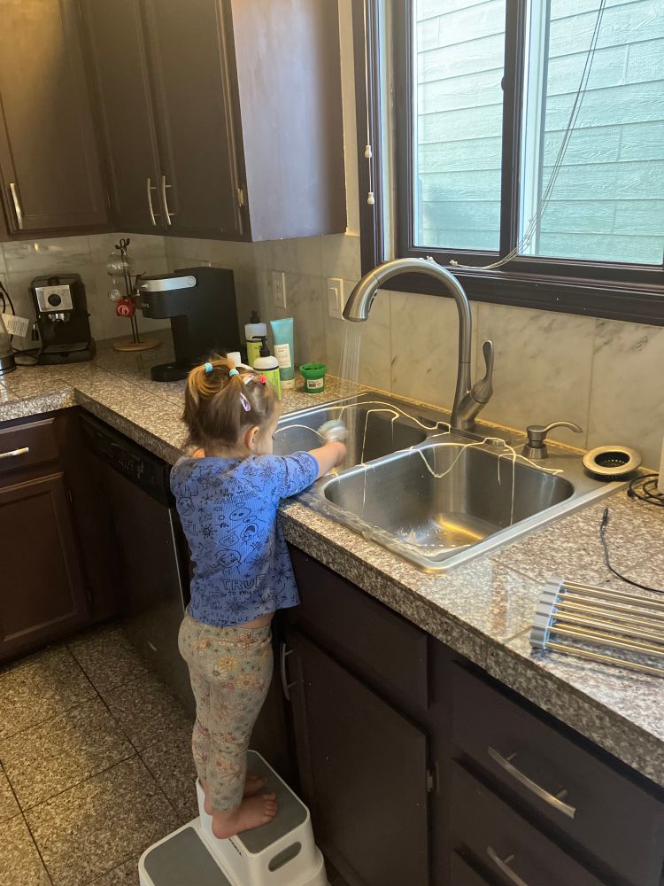 Toddler with a blue shirt and ponytail standing on a stool in the kitchen, scrubbing the sink. 