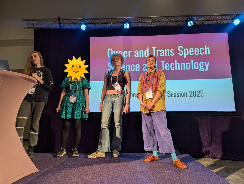 Four well-dressed people, one speaking, stand on a stage in front of a large screen displaying the text "Queer and Trans Speech Science and Technology: Interspeech Special Session 2025"