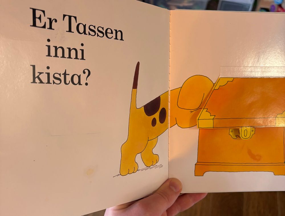 Bilde fra boken «hvor er Tassen?», med teksten «Er Tassen inni kista?», og bilde av en hund som kikker oppi en kiste.