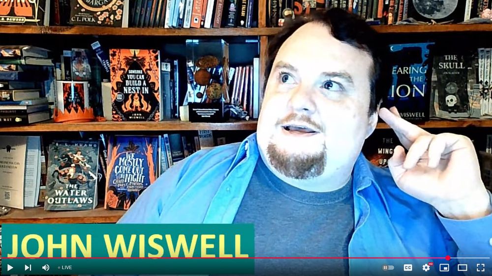 John looking bewildered in front of a shelf of books. The name JOHN WISWELL is on the bottom of the screen.