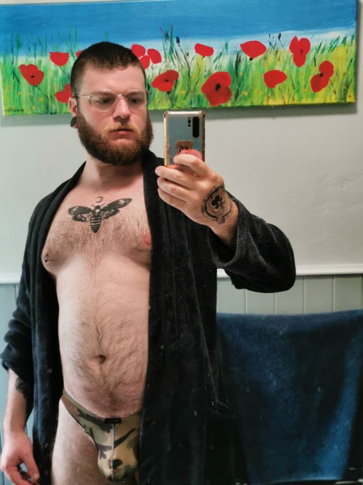 Bathroom mirror selfie of me in a pair of daddy looking glasses in a bathroom that is left open to show a camo print thong with the imprint of a chastity cage visible 