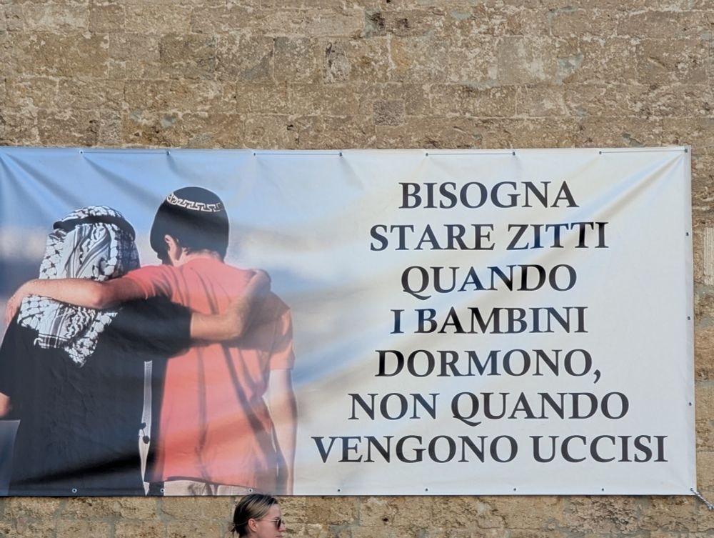 Banner hanging outside a church in San Gimignano, Italy depicting a child wearing a keffiyeh and another wearing a kippah with their arms around each other. Next to them is text in Italian that translates as "You should be quiet when children are sleeping, not when they're being killed."