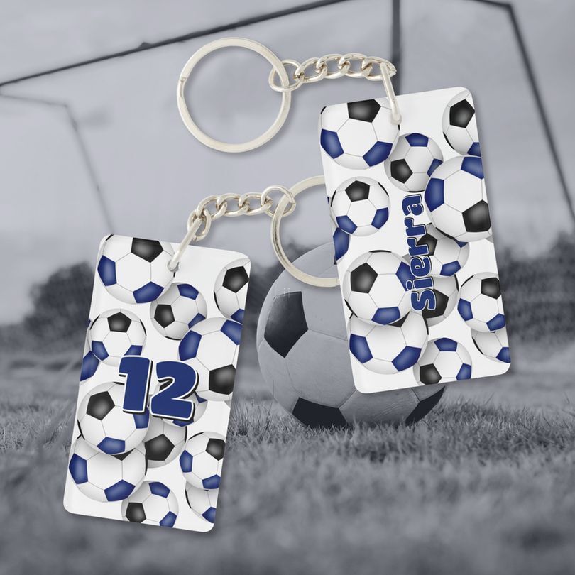 Mockup image of a soccer themed keychain, front & back views. Each side of the keychain features the image of a cluster blue, black & white soccer balls. The keychain is personalized with the athlete’s name on the front and jersey number on the back. Background scene for the mockup is a soccer field with a soccer ball in the foreground and soccer goal in the distance in black & white.