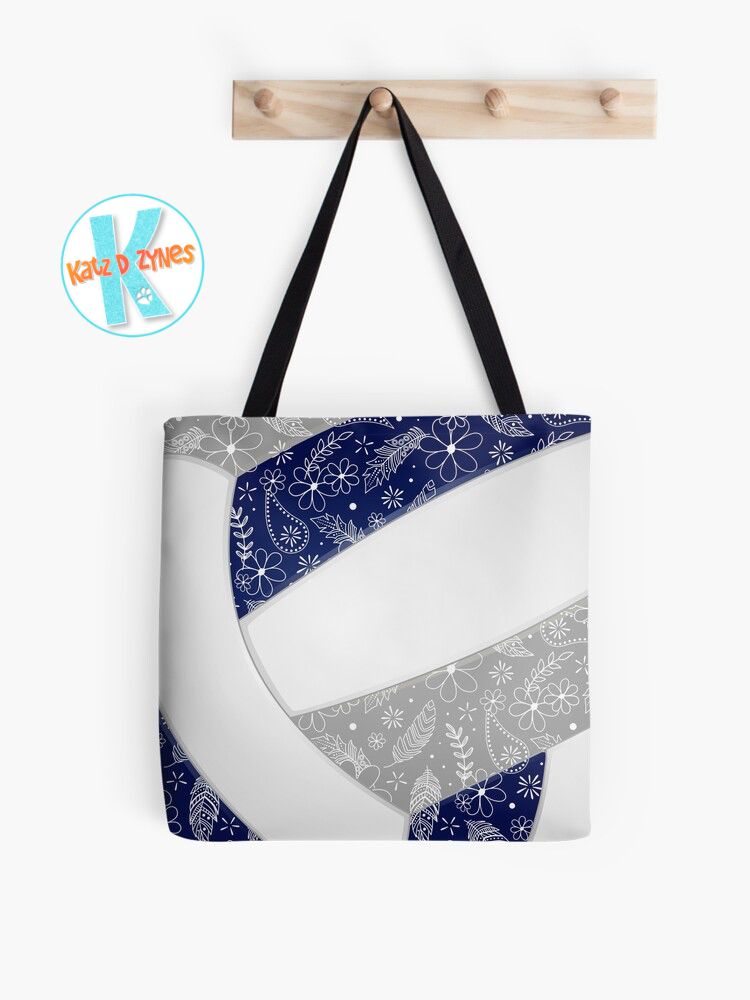 Mockup display of a volleyball-themed tote bag with black straps hanging on a wooden peg rack against a white wall. The bag is fully covered with the image of an abstractly oversized volleyball that has navy blue, gray and white panels. A delicate boho doodle pattern of paisleys, feathers and flowers in white overlies the volleyball’s navy & gray panels.