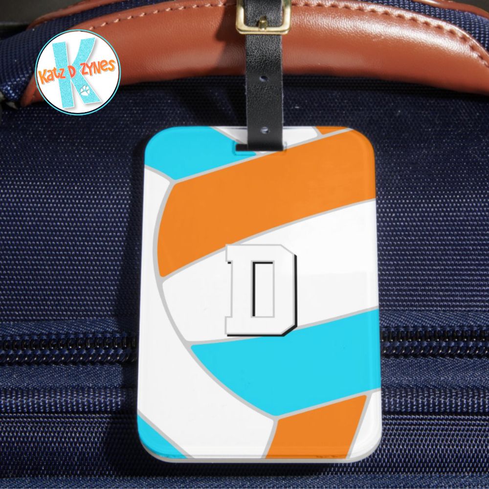Mockup image of a volleyball themed luggage tag, displayed hanging from the leather handle of a blue fabric travel bag. The image of an abstractly oversized volleyball with bright blue, orange & white panels fully covers the tag. The tag is personalized with an athlete’s monogram, the letter D.