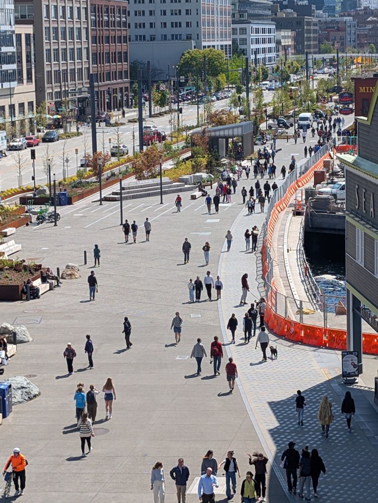 The waterfront promenade in front of the Seattle Aquarium showing the wide walkway and 2 way fully protected bike lane. Sunny day with many people strolling along the walkway.