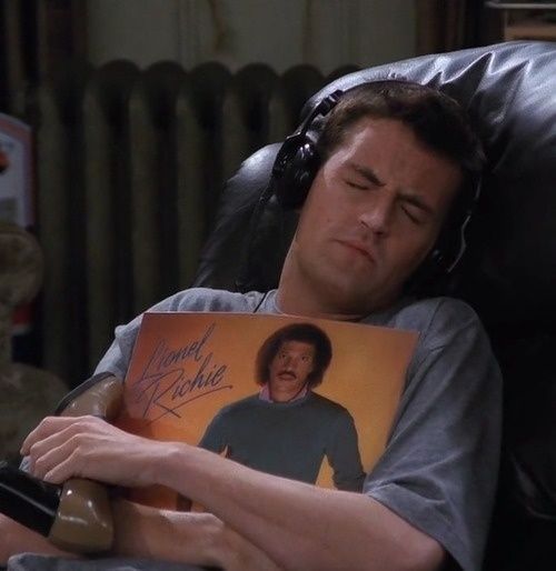 Chandler abraçando o álbum do Lionel Richie usando fones de ouvido e segurando um dos sapatos da Janice 