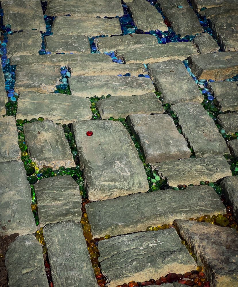 Patio pavers in a alternating square pattern. In between each paver are colored glass marbles, creating a gradient, rainbow across the path.
