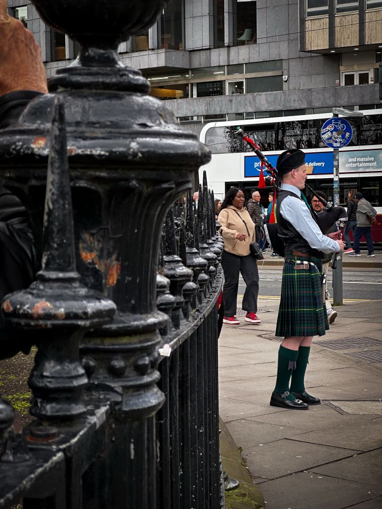 A musician in full Scots regalia plays bagpipes on busy Princes Street corner. There is an iron fence in the foreground.