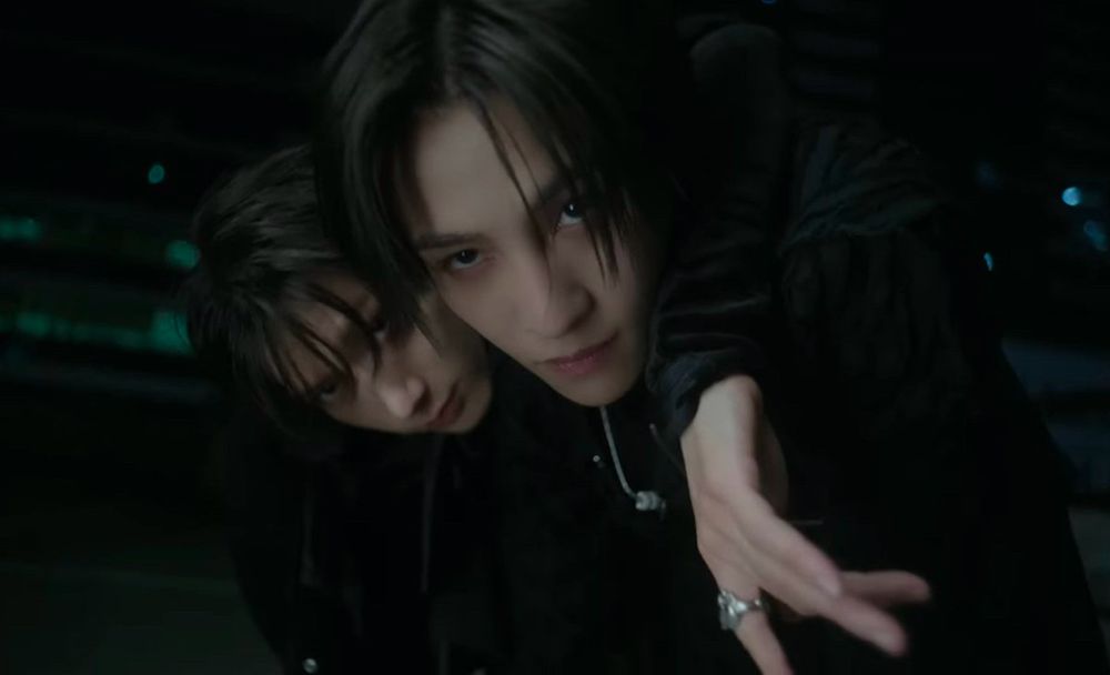 Ten with his arm around Yangyang's neck, both leaning towards the camera. They have black hair and are wearing black and the background is dark too.