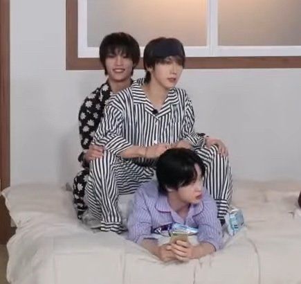 Yangyang and Ten sitting on Kun's back, who's lying down on his belly on a mattress. They're all in pyjamas, Yangyang wearing a black one with daisies, Ten a black and white striped one, and Kun a light blue one. They all have black hair. Yangyang is grinning widely.