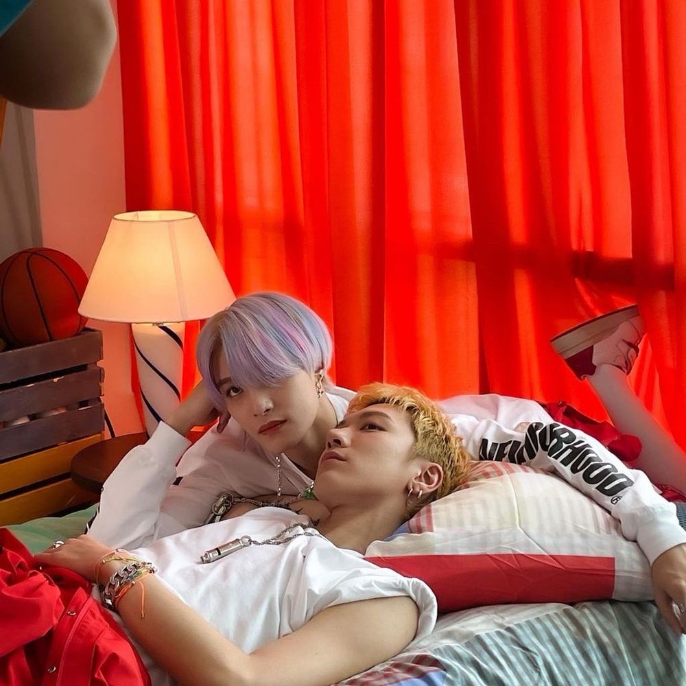 Yangyang and Ten at the set of low low. 
They're both on a bed, Yangyang on his stomach and Ten on his back, their heads next to each other. They're both wearing white shirts and Ten is wearing red trousers and short yellow hair. Yangyang's hair is lilac and almost a bowl cut. 
Behind them are red curtains, a lamp and a box with a basketball in it.