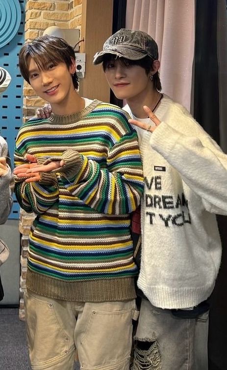 Ten and Yangyang posing for a group picture, smiling.
Yangyang has his hand on Ten's shoulder and is showing a peace sign. Ten is gesturing at their album
Ten is in a striped sweater in green, yellow, brown and blue and Yangyang in a white sweater with black text on it, a black cap and ripped jeans.