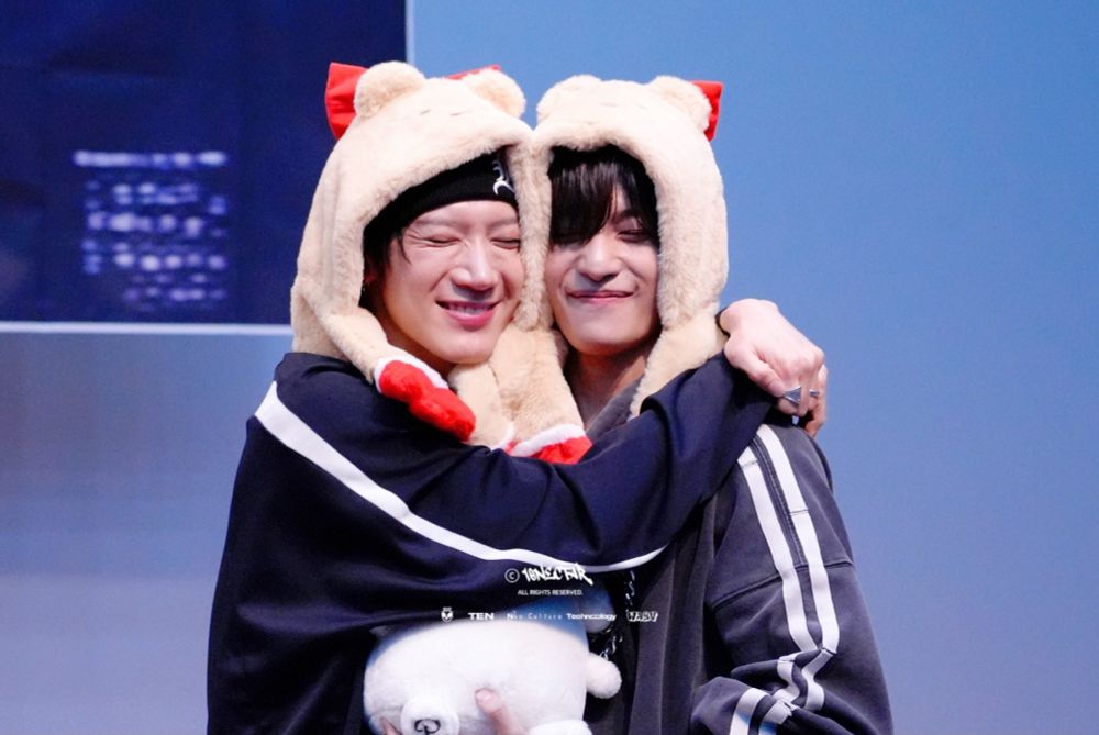 Ten and Yangyang hugging with their cheeks pressed against each other. They have black hair and are both wearing dark blue sports jackets and beige, fluffy hats with round ears and faces. The hats have long arms with red gloves too. Theyre both smiling and squeezing their eyes shut and Yangyang is holding a plushie.